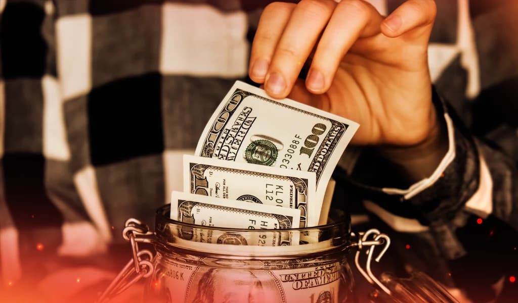 Close-up of a hand placing hundred-dollar bills into a glass jar, symbolizing saving or poker winnings.