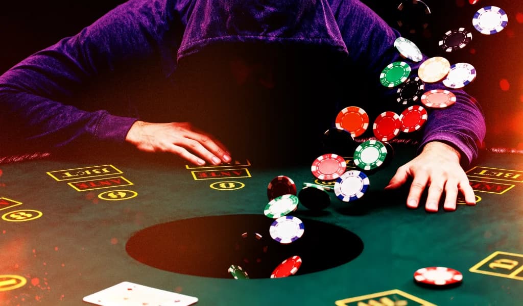 Poker player with a hoodie over his head leaning over a table as colorful chips fall into a dark hole at the center of the table.