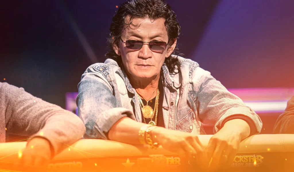 Scotty Nguyen, wearing sunglasses and a denim jacket, leaning over a poker table with gold jewellery and serious focus.