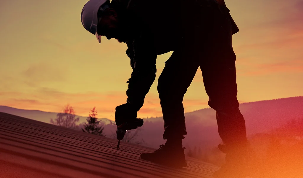 Silhouette of a roofer drilling on a roof during sunset, with a mountain backdrop and colourful sky.