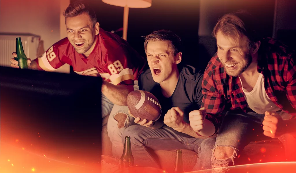 Three men cheer intensely while watching a football game on TV, one holding a football and another wearing a red jersey. Warm lighting adds to the excitement.