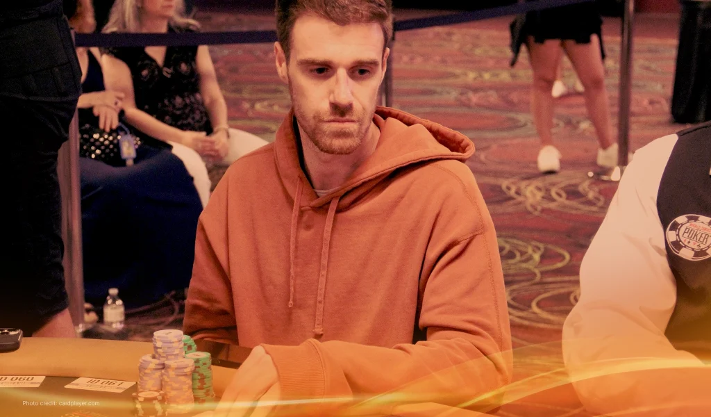 Adrian Attenborough, wearing an orange hoodie, sits at a poker table with stacks of chips, looking focused.