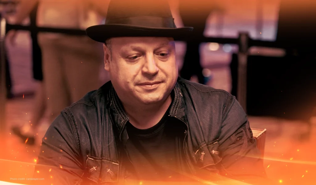 Jeff Lisandro, wearing a black hat and jacket, smiles slightly while sitting at a poker table in a tournament setting.