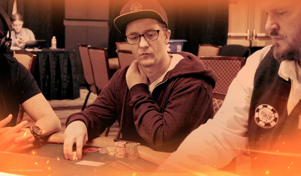 Kahle Burns, wearing a cap and glasses, plays poker at a tournament table, focused on his hand while holding chips.