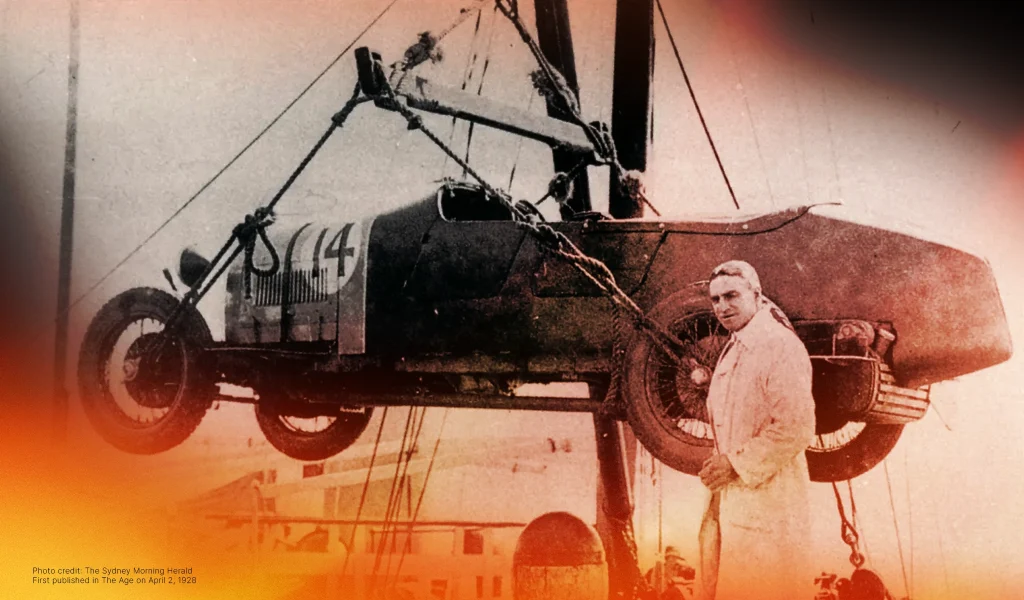 Vintage car with the number 14 being hoisted by a crane, with a man in a white coat standing nearby.