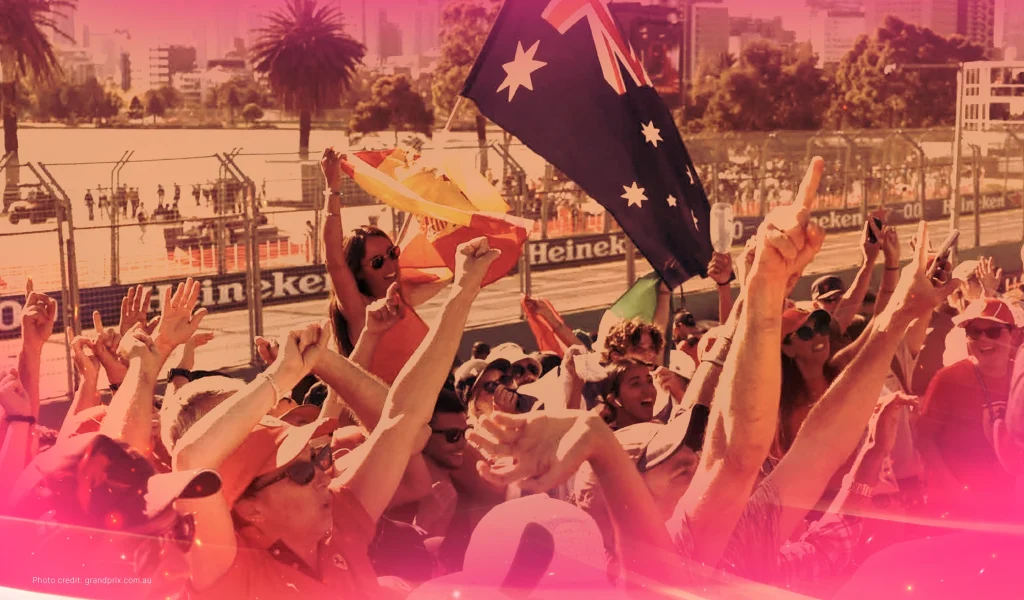 Excited crowd at the Australian Grand Prix, waving flags, including an Australian flag, with city buildings and palm trees in the background.