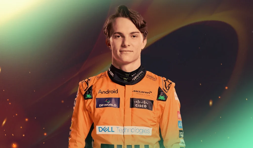 Young race car driver, Oscar Piastri, in an orange McLaren racing suit stands against a dark background.