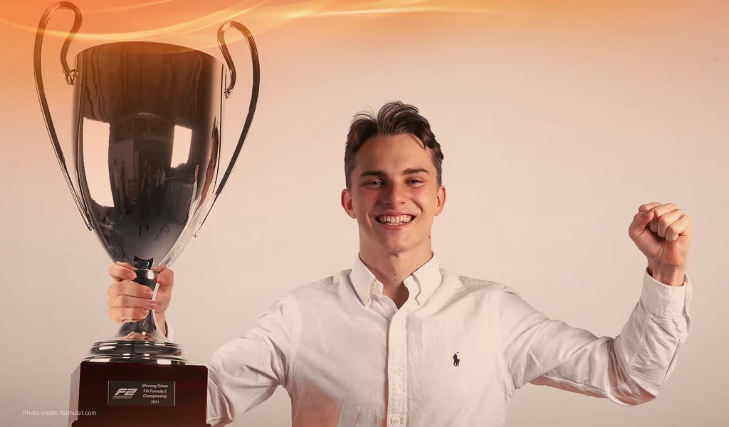 Smiling young Oscar Piastri in a white shirt holds up a large silver trophy in victory.