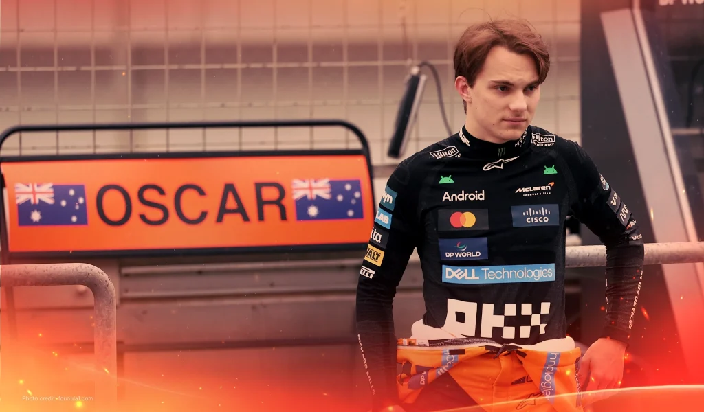 Race car driver in a black McLaren suit stands near a sign displaying "Oscar" with Australian flags.