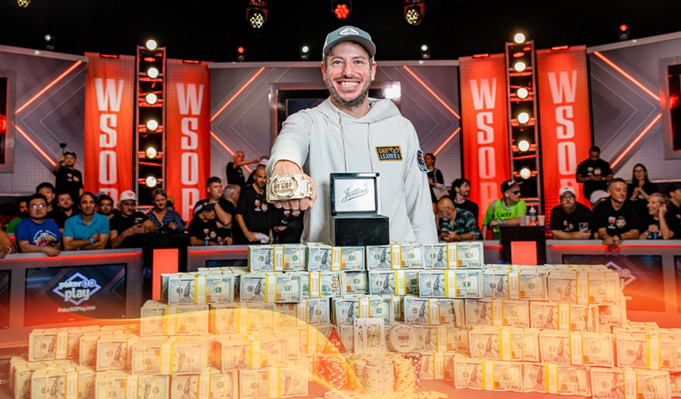 Daniel Weinman, winner of the World Series of Poker 2023 holding a championship bracelet, standing behind stacks of cash, with a cheering audience in the background.