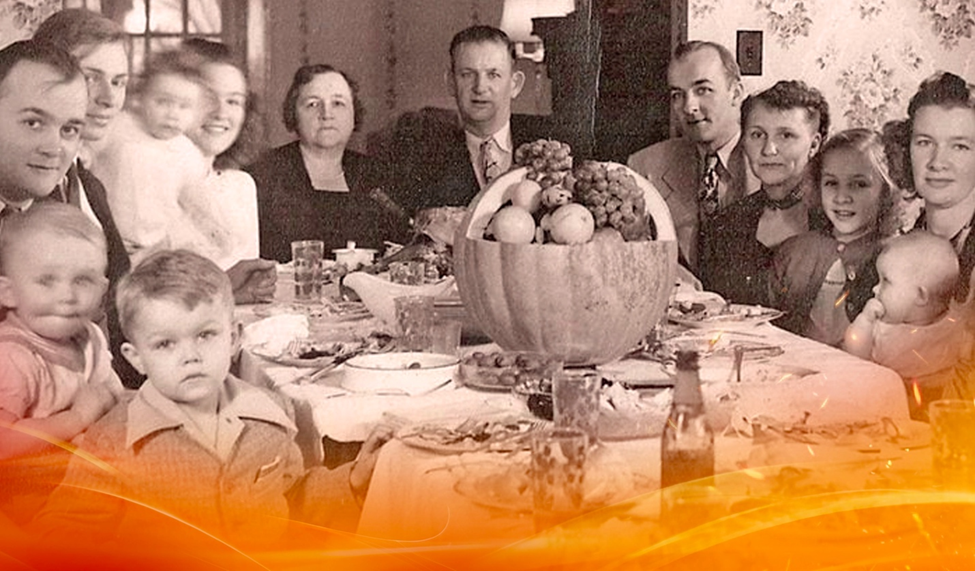 A black-and-white photo of a family gathered around a Thanksgiving table with food and a pumpkin centerpiece.