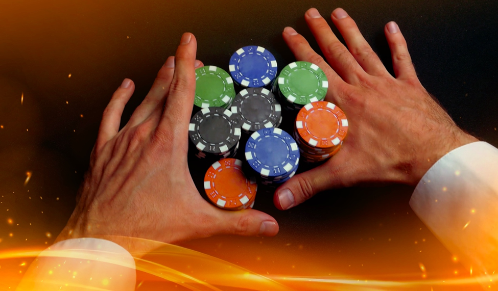 A pair of hands surrounds a large stack of colorful poker chips, including green, blue, black, and orange, on a dark table with glowing orange accents.